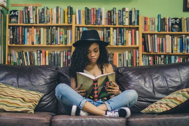 A younger girl wearing a brimmed hat sits on a couch reading a book, with a fairly large amount of shelved books behind her against a wall.