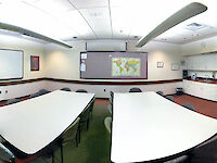 An overview of the entirety of the room: Two tables with 8 chairs each, a well-lit room with whiteboard and large bulletin board, and small kitchenette.