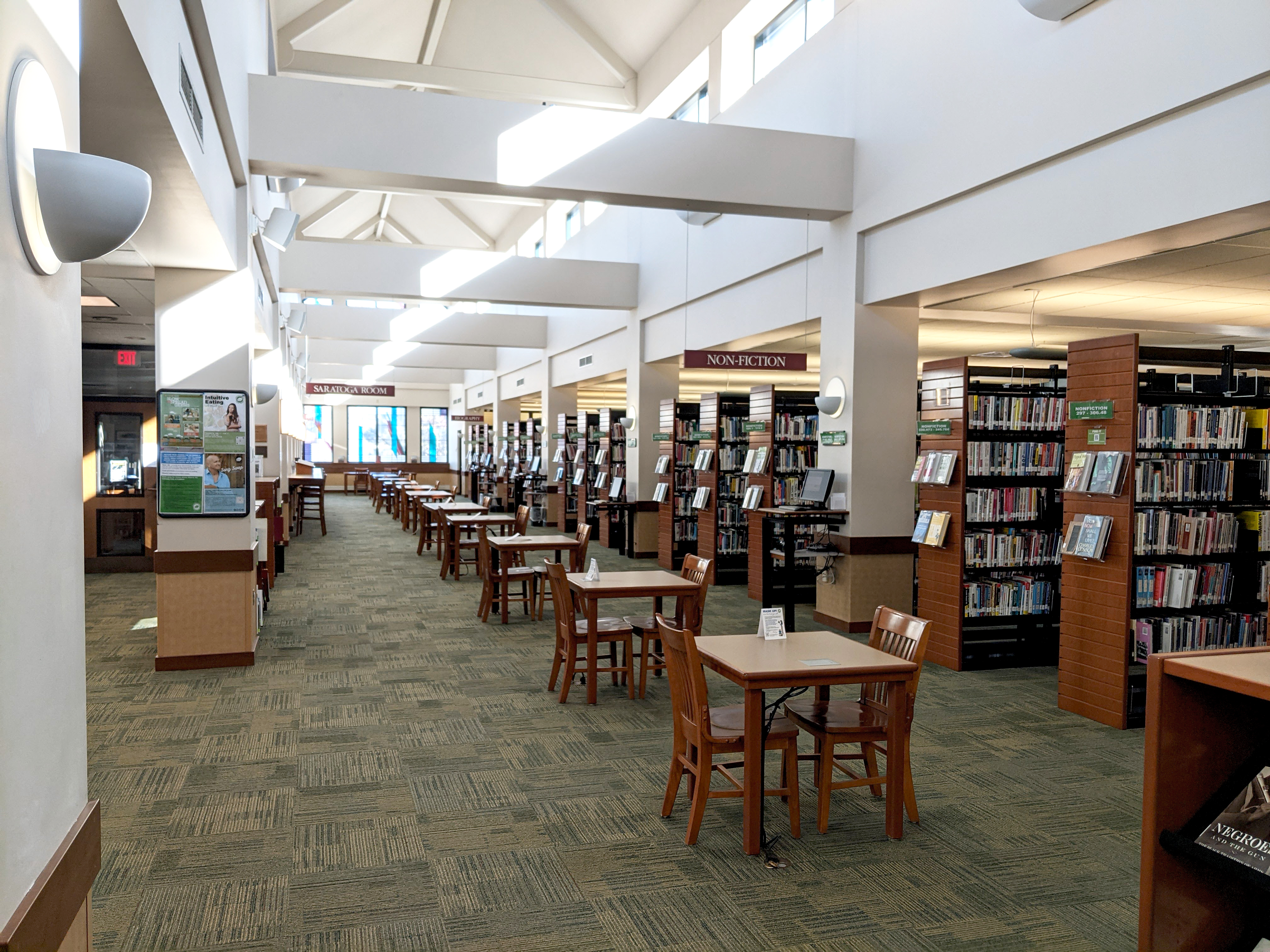 A long floorspace with many evenly spaced tables set out in front of a long row of book shelving and high, open ceilings with natural light.