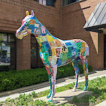 Thumbnail: A decorated horse statue with various painting designs covering its body, standing in the center of a garden bed area next to a walkway.