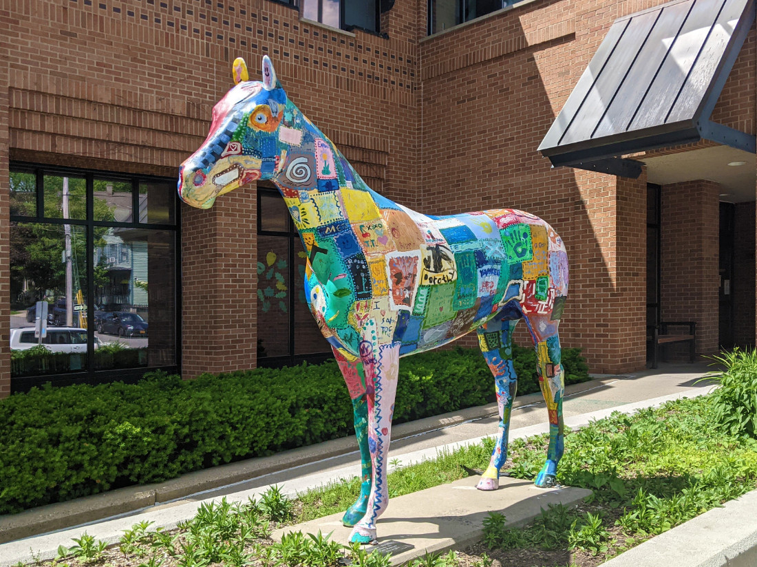 A decorated horse statue with various painting designs covering its body, standing in the center of a garden bed area next to a walkway.