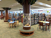 Tree-like columns, decorative wooden chairs, and shelves with animal-inspired endcap artwork withmany books on shelves.