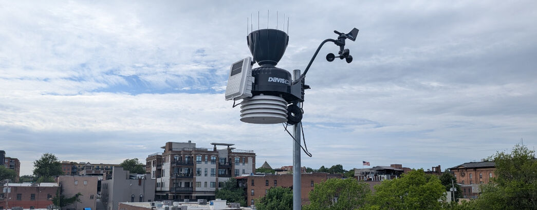 A Davis Weather station mounted atop the library building roof, overlooking the backdrop of the buildings on Broadway in downtown Saratoga Springs.