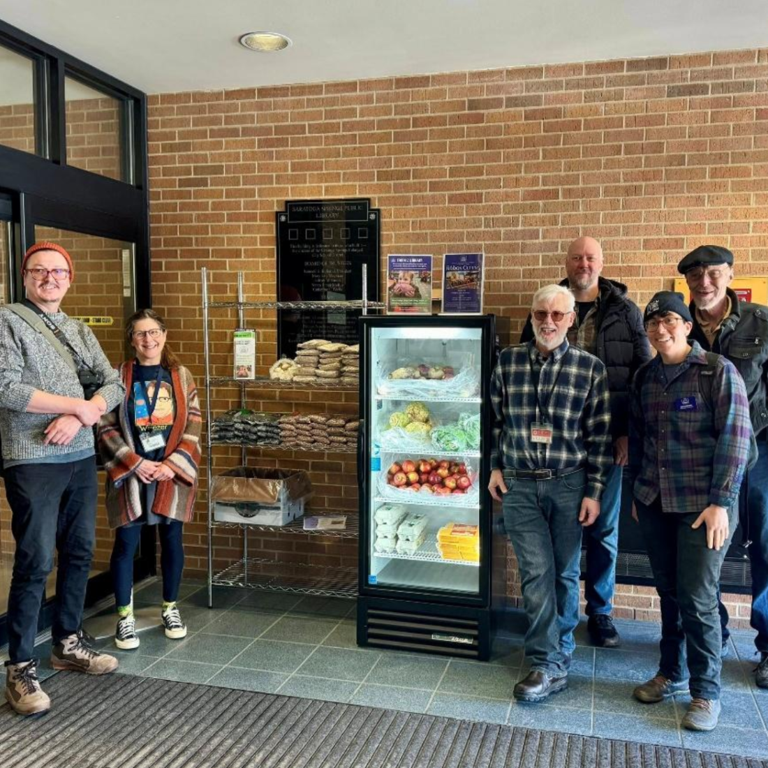 Six people stand around refrigerated and dry goods shelves inside a foyer.