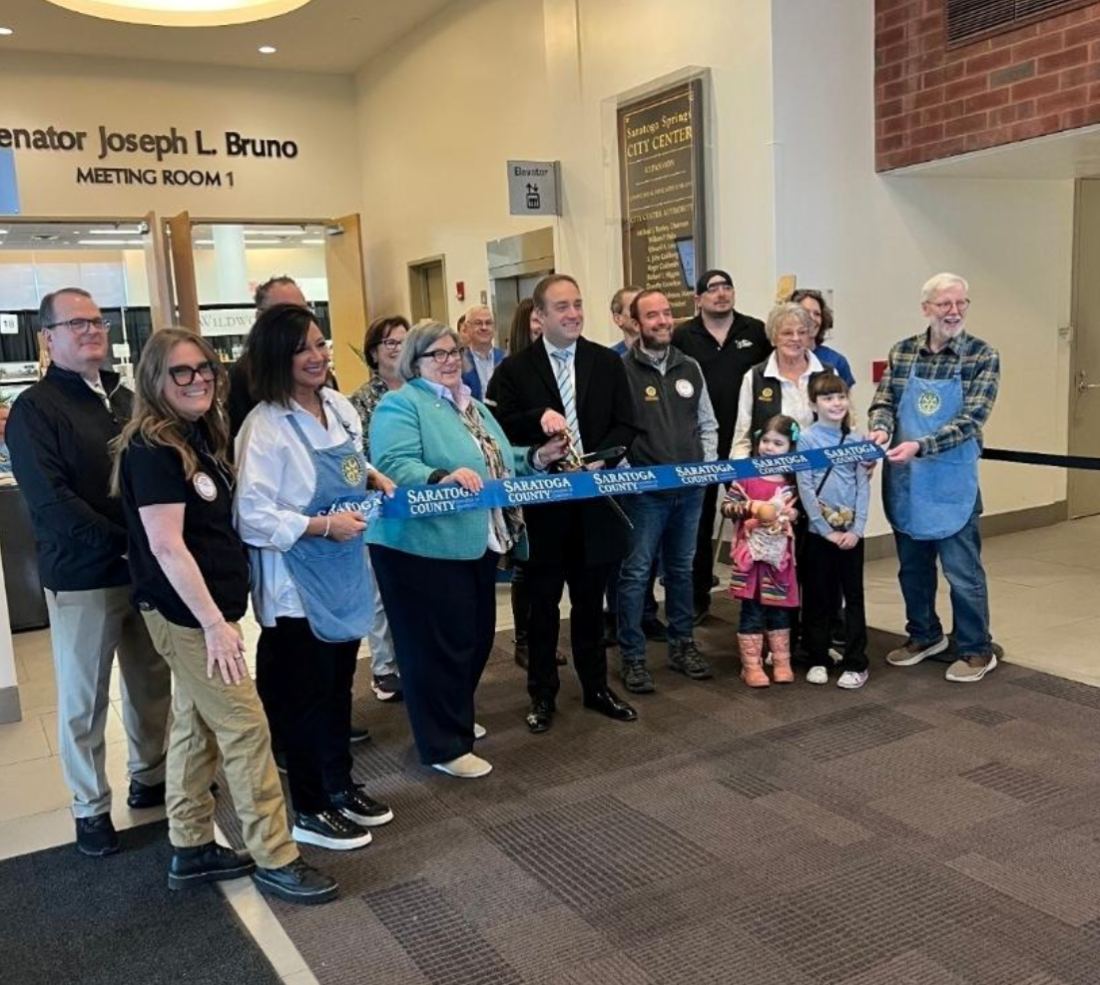 17 people stand behind a ribbon with scissors open and ready to cut in celebration.