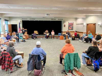 A group of people seated in chairs, set in a spacious and large round circle, participate in a drum-circle of percussion.