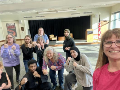 Smiling women, hands clenched and fists up, strike a pose as they huddle around the instructor.