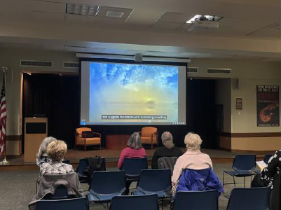 The audience waits for the program to begin as a beautiful photograph of the sunset over water is displayed on the screen.