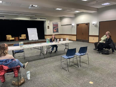 Two long tables, connected lengthwise, are adorned with informational materials and sample Ecobricks, while attendees listen to the presenter provide details.