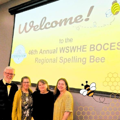 Library Director, Assistant Director, Head of Youth Services, and Head of Adult Services staff stand in front of the regional spelling bee’s presentation screen’s welcome message.