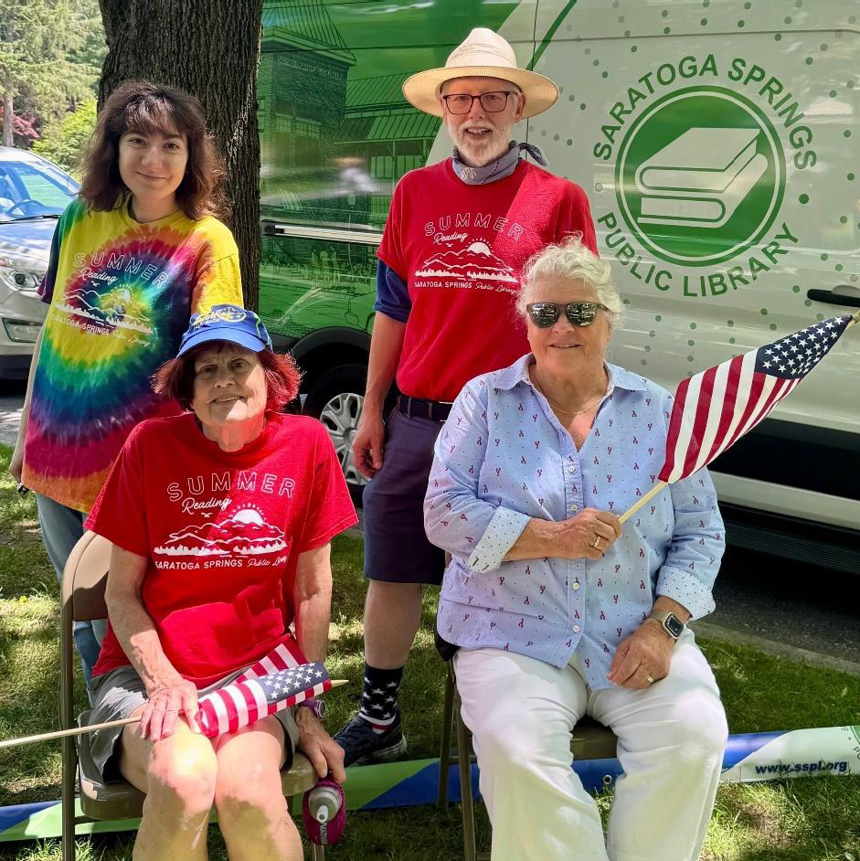 Four people pose in front of the library van, two seated and two standing, representing the library and Flag Day with appropriately colored garments.