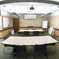 Three tables with 8 chairs each equidistant within the room, with a whiteboard and projector screen in the back of the room, bulletin board to the right wall, and a kitchenette to the left.