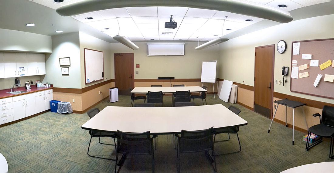 Three tables with 8 chairs each equidistant within the room, with a whiteboard and projector screen in the back of the room, bulletin board to the right wall, and a kitchenette to the left.