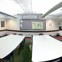 An overview of the entirety of the room: Two tables with 8 chairs each, a well-lit room with whiteboard and large bulletin board, and small kitchenette.