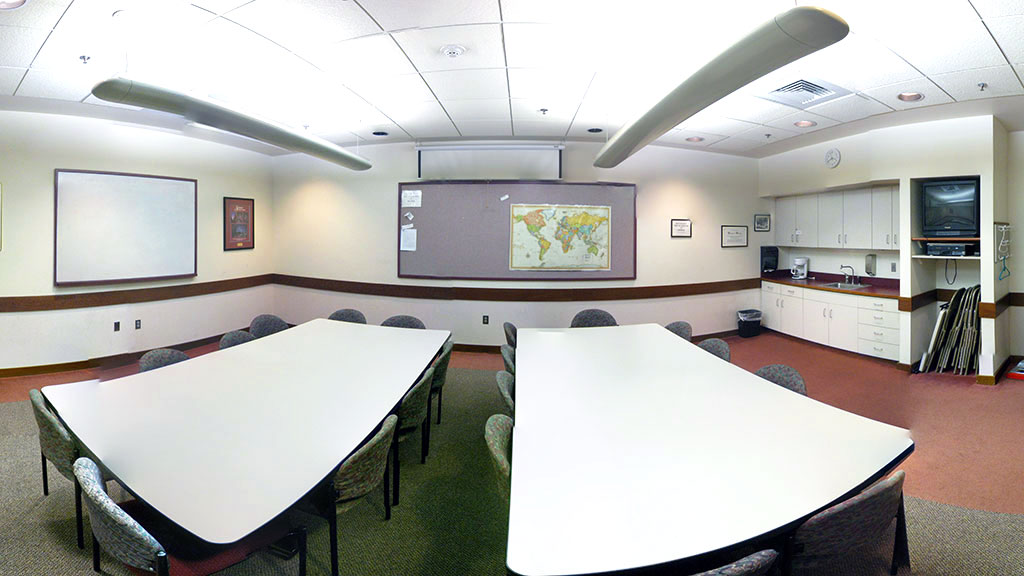 An overview of the entirety of the room: Two tables with 8 chairs each, a well-lit room with whiteboard and large bulletin board, and small kitchenette.