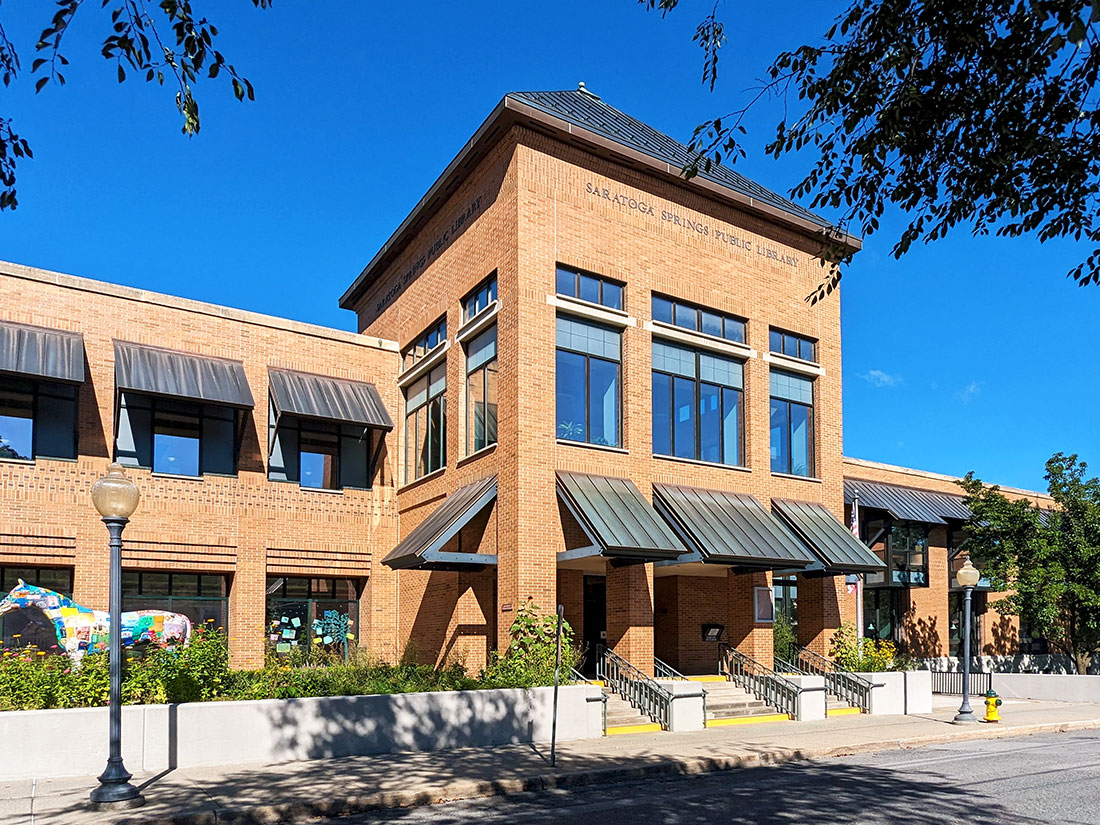 Shadows from overhead trees line the street in front of the main entrance to the Saratoga Springs Public Library on a day with a clear, blue sky.