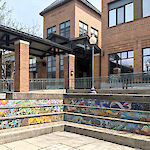 Thumbnail: A layered, step-like tile mosaic behind a brick paver patio with garden beds outside and the library’s Putnam Street entryway and brick tower in the background.