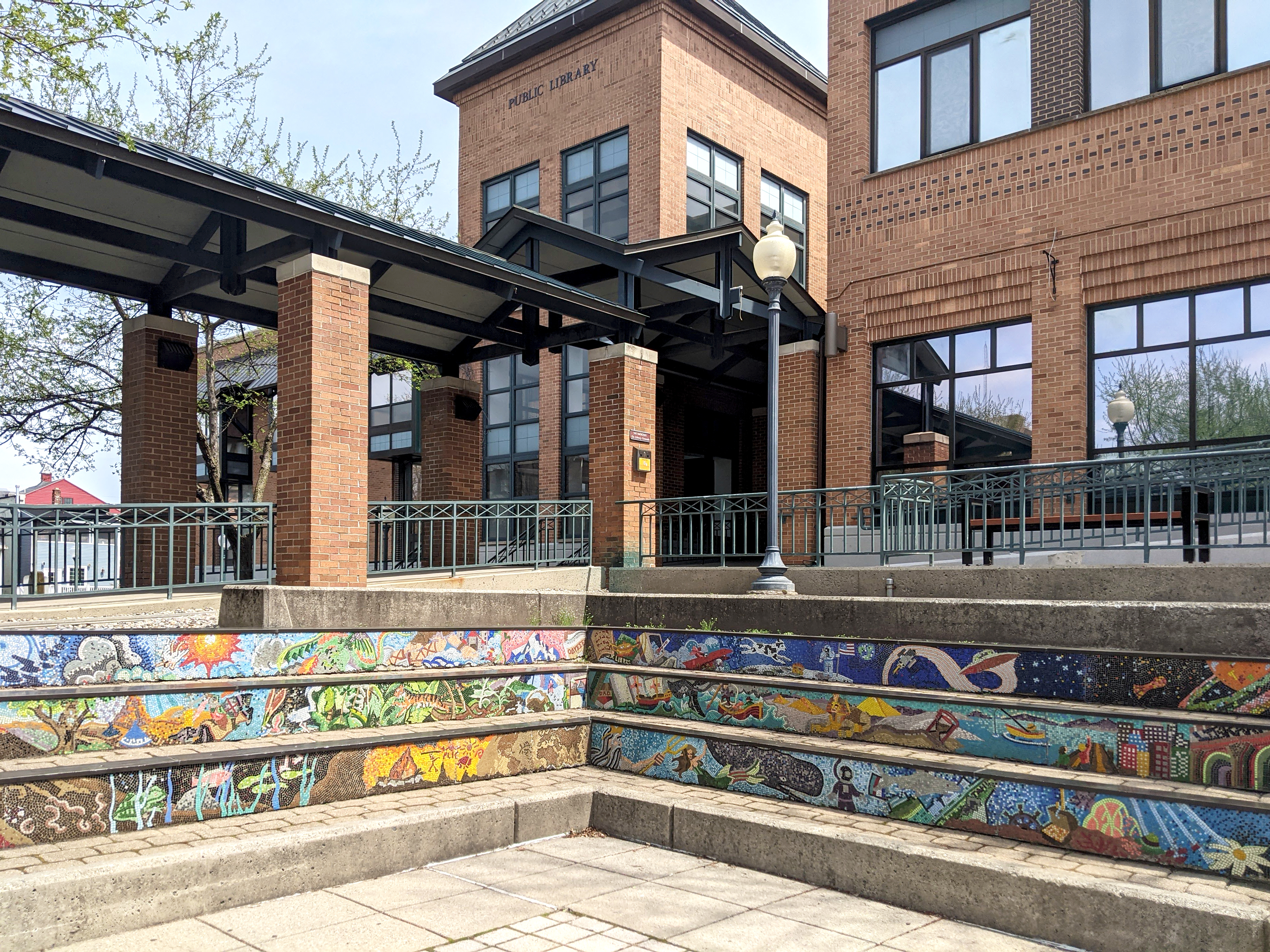 A layered, step-like tile mosaic behind a brick paver patio with garden beds outside and the library’s Putnam Street entryway and brick tower in the background.
