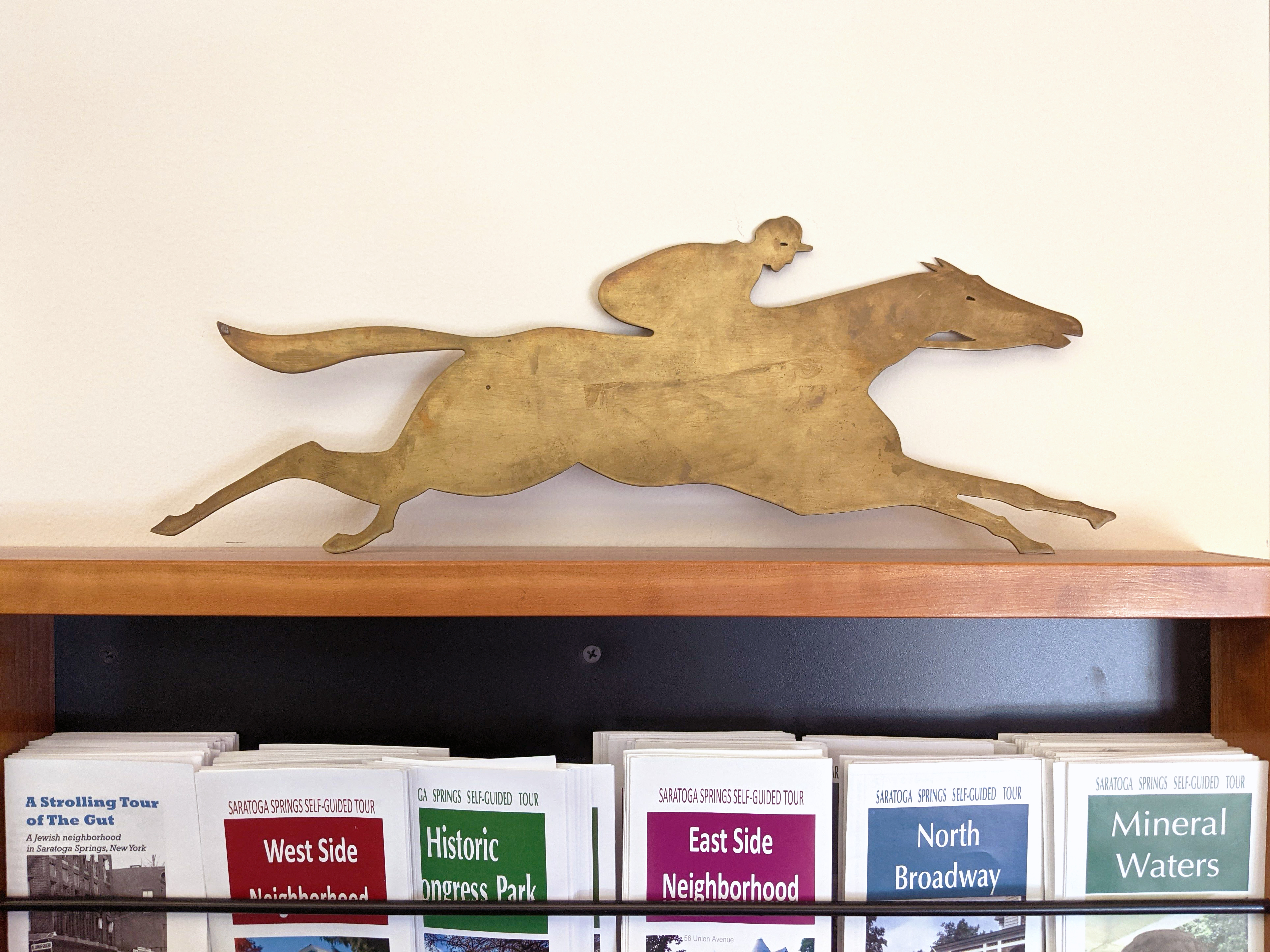 Bronze-like placque of a jockey riding a thoroughbred to grab attention, laid atop a display case to provide trifold printed pamphlets for self-guided tours.
