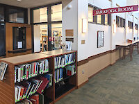 A door partially hidden by a waist high shelf of books with a sign to the right hanging from the ceiling to denote the entrance to the Saratoga (History) Room.