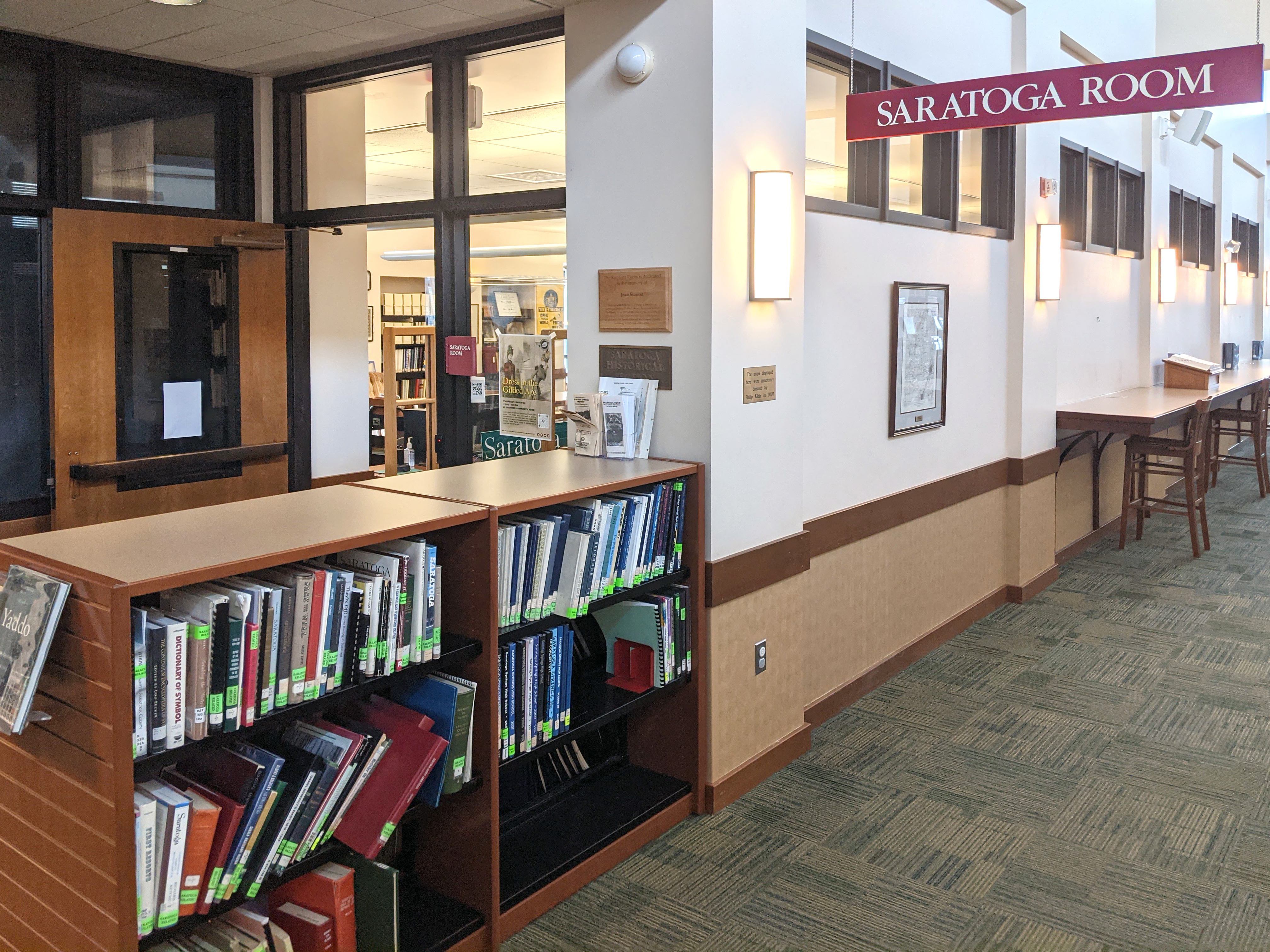 A door partially hidden by a waist high shelf of books with a sign to the right hanging from the ceiling to denote the entrance to the Saratoga (History) Room.