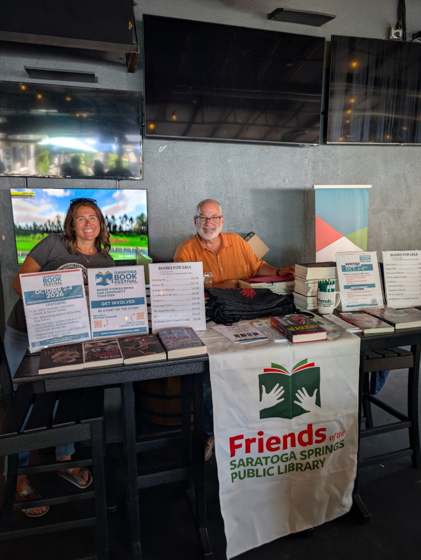 A woman and a man smiling while standing behind a tall table. There’s a banner that reads “Friends of the Saratoga springs Public Library.” Books for sale are on the table..