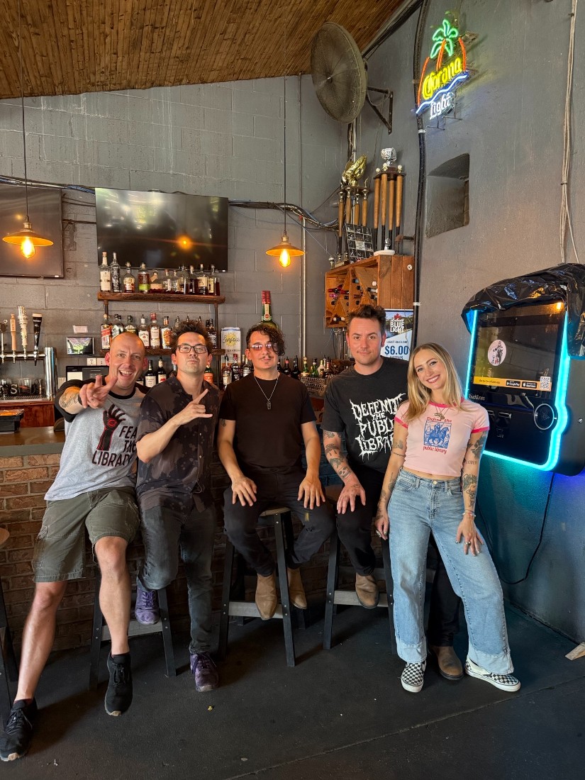 Five people seated at a bar, facing away from the bar, smiling and pointing to a person taking the photo.