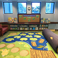 A play area with chairs, corkboard bulletin area, padded log seat, and decorative nature-themed carpet.