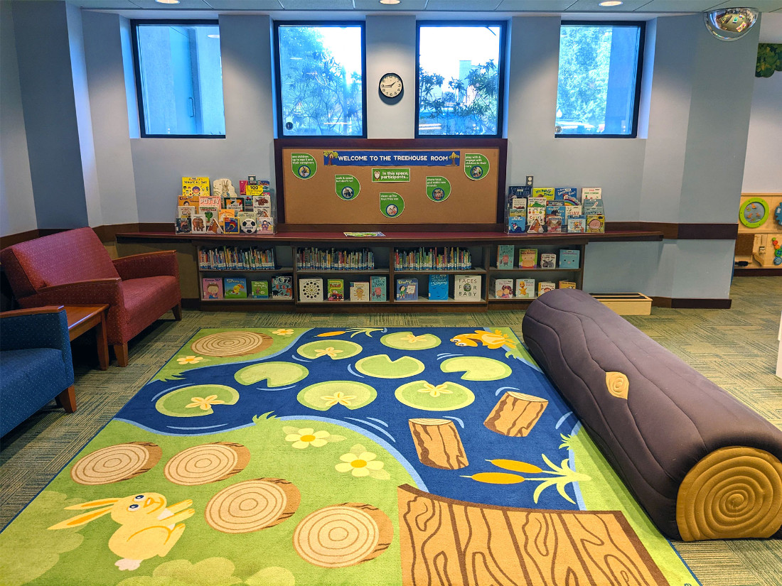 A play area with chairs, corkboard bulletin area, padded log seat, and decorative nature-themed carpet.