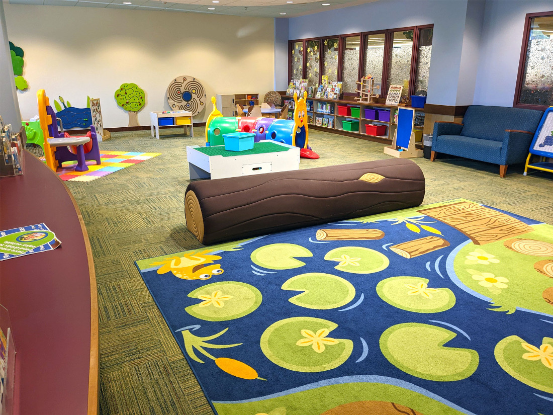 A play place with a padded log bench, lily pad carpet, and scattered play things.