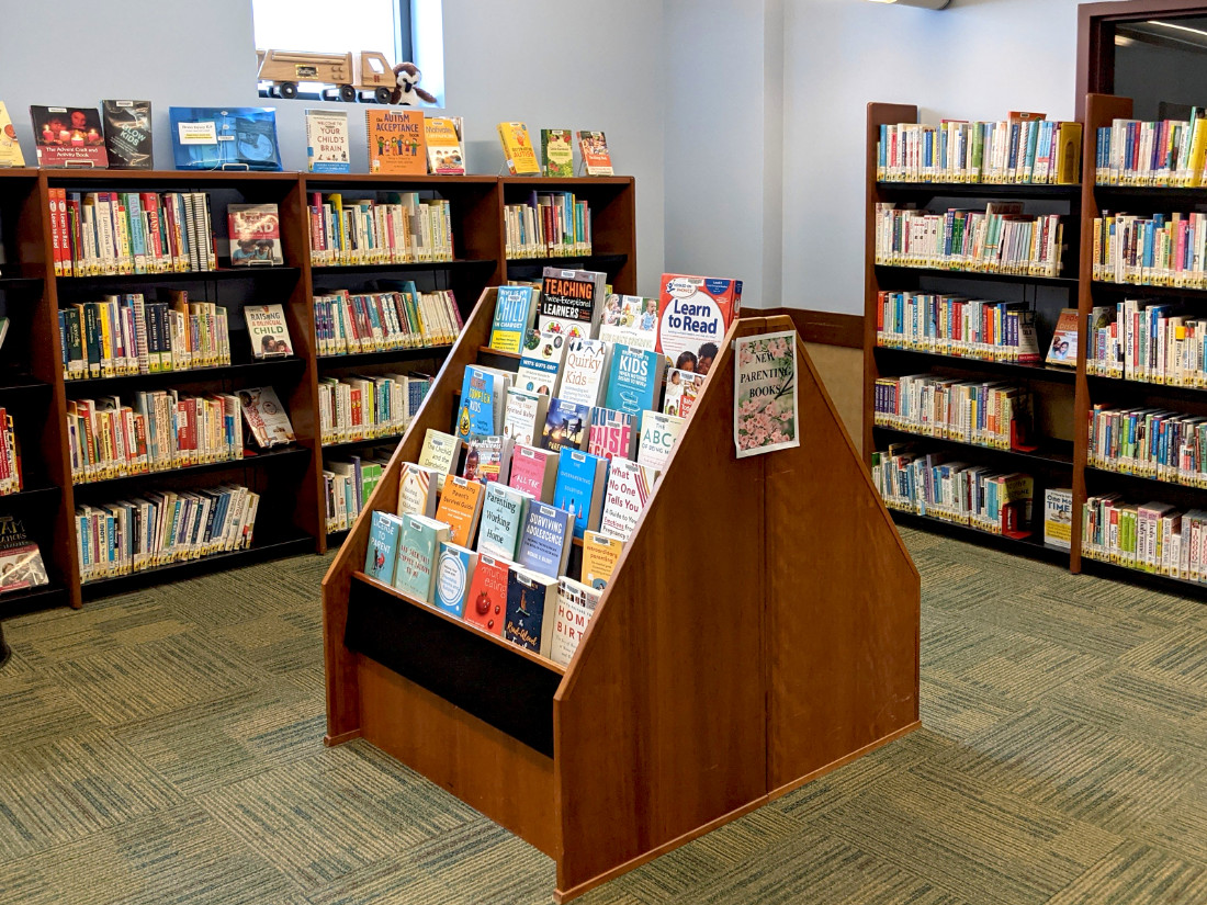 A small floor standing stack of selected items in front of two large shelves against corner walls full of books.