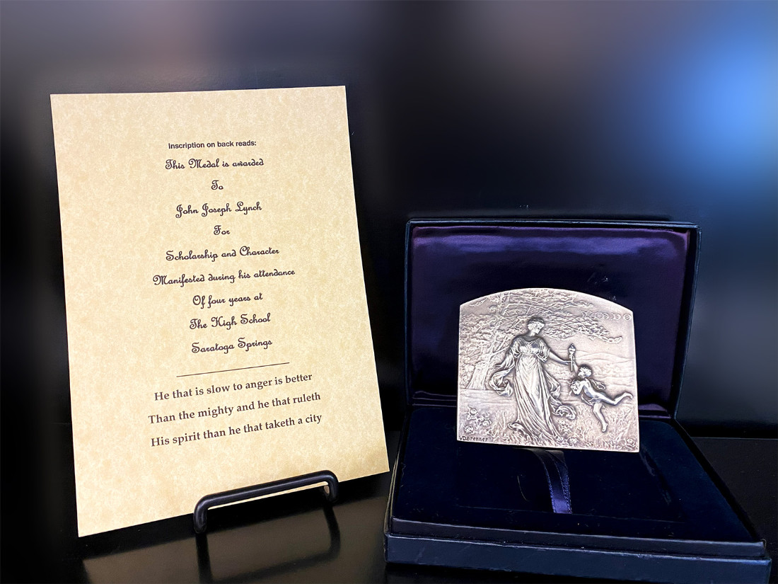 A top-rounded rectangular medal stands in a decorative, felted box. To its left is a ceremonial certificate on parchment declaring the award to the recipient.