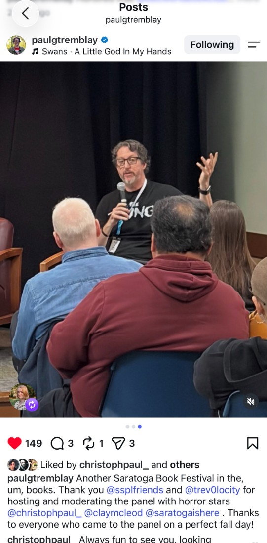 Horror author, Paul Tremblay, speaks into a handheld microphone while seated on a stage in front of a black curtain. Several audience members are seated in the foreground, facing the speaker.