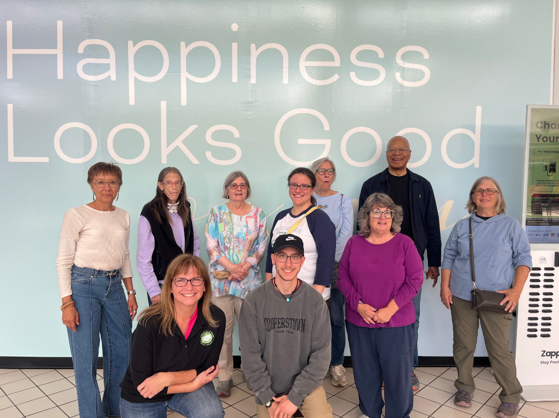 A group of 10 adults are posed in front of a wall that says “Happiness Looks Good”
