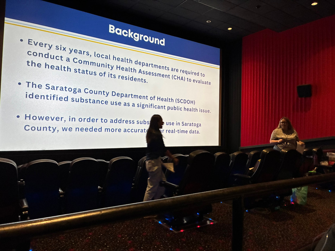 Two women are standing in front of a movie screen talking about the history of Narcan training in Saratoga County