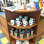 Thumbnail: Decorated cloth shopping bags, ceramic and travel mugs fill an end-shelf’s display area for purchase.