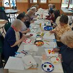 Thumbnail: long white table with people sitting on either side.  windows and wooden doors in back of room.  people painting at table