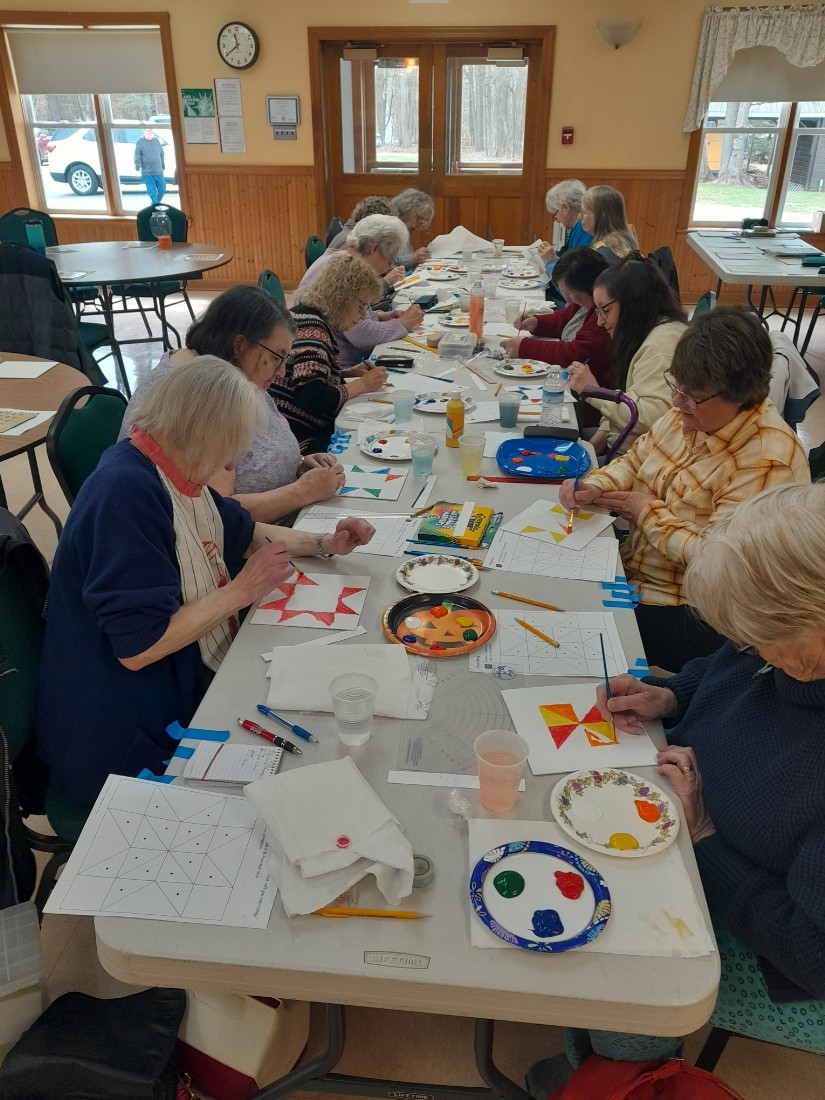 long white table with people sitting on either side.  windows and wooden doors in back of room.  people painting at table