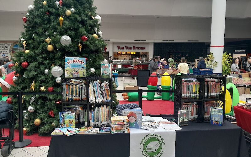 Inside Wilton Mall food court. long black table with white table runner.  books stacked in crates on table.  Christmas tree behind table.  People in the background behind the Christmas tree.
