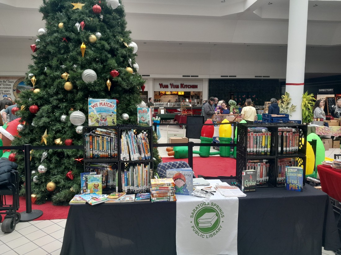 Inside Wilton Mall food court. long black table with white table runner.  books stacked in crates on table.  Christmas tree behind table.  People in the background behind the Christmas tree.