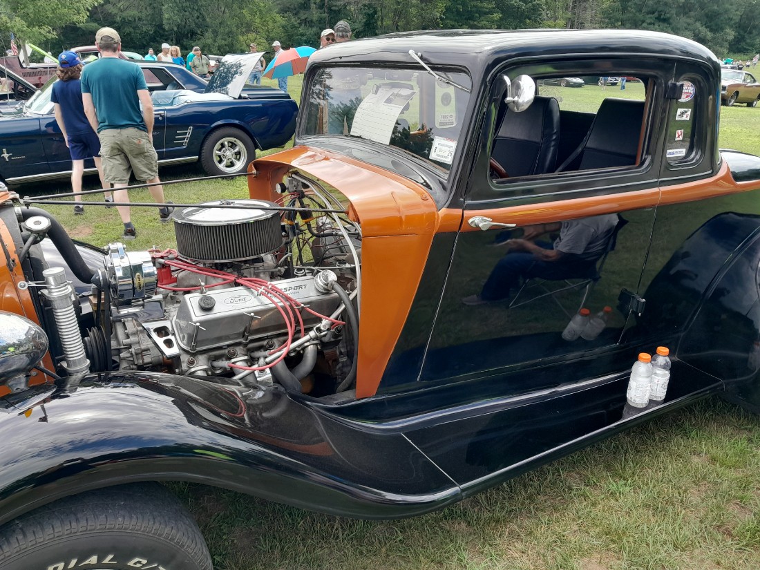 vintage car with black top and runners. copper colored engine cover. exposed engine. car located in park with additional cars and people in the background