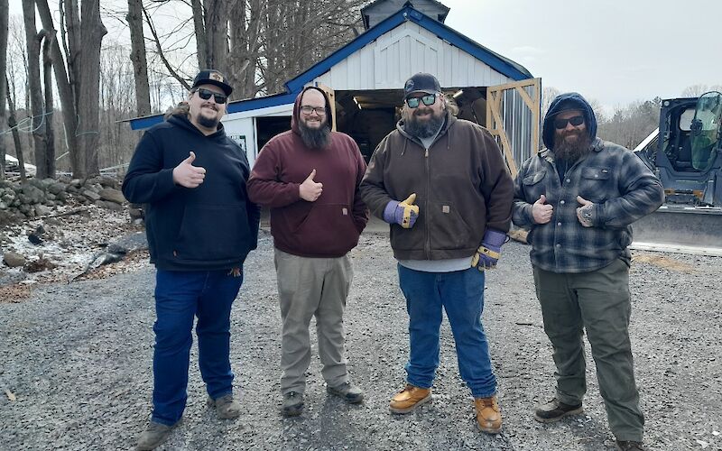 four men with a thumbs up in front of a maple sugar shack.  Men wearing blue, red, brown and flannel sweatshirts.  Trees and tractor in the background.