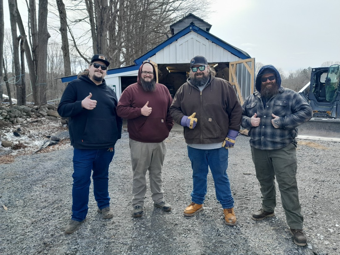 four men with a thumbs up in front of a maple sugar shack.  Men wearing blue, red, brown and flannel sweatshirts.  Trees and tractor in the background.