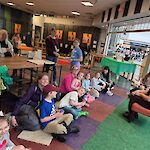 Thumbnail: woman wearing black and green in front of room sitting on a red chair. Group of children and adults sitting on carpet squares facing woman.