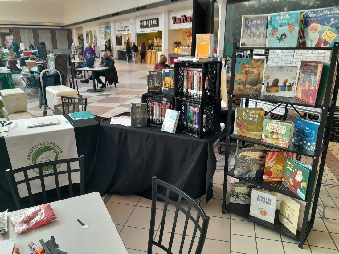 5 shelf metal bookcase on right with picture books displayed on it. table with a black tablecloth to the left of the bookcase with additional books and a library table runner.